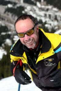 Un moniteur de ski souriant portant des lunettes de soleil et une veste de ski jaune et noire sur une pente enneigée.